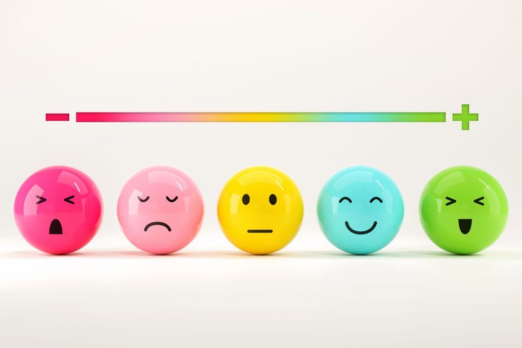 Colorful round emoji faces representing a range of emotions from anger to joy, arranged beneath a gradient scale from negative to positive—highlighting how emotions are often labeled and ranked rather than understood as equally valid.