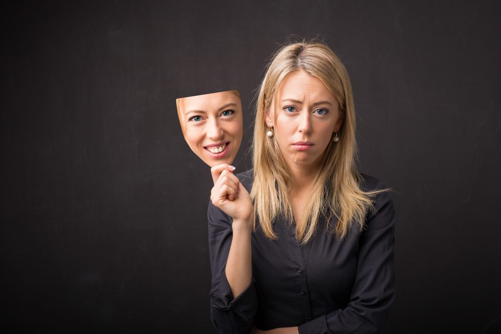 Woman holding a smiling mask in front of her face while looking sad, symbolizing the emotional conflict and hidden struggle of people-pleasing in adulthood.