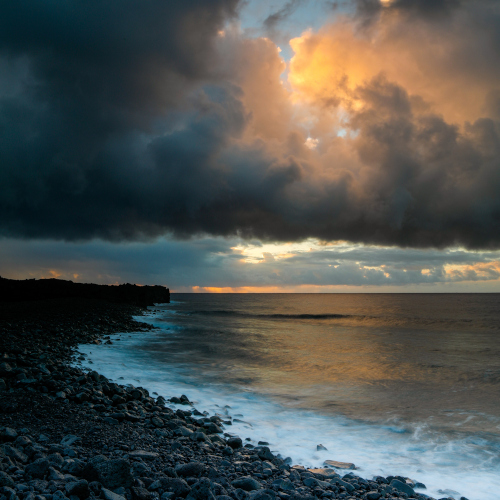 A dark shoreline under heavy clouds opening to warm light over calm water, reflecting the movement from survival toward safety and steadiness.