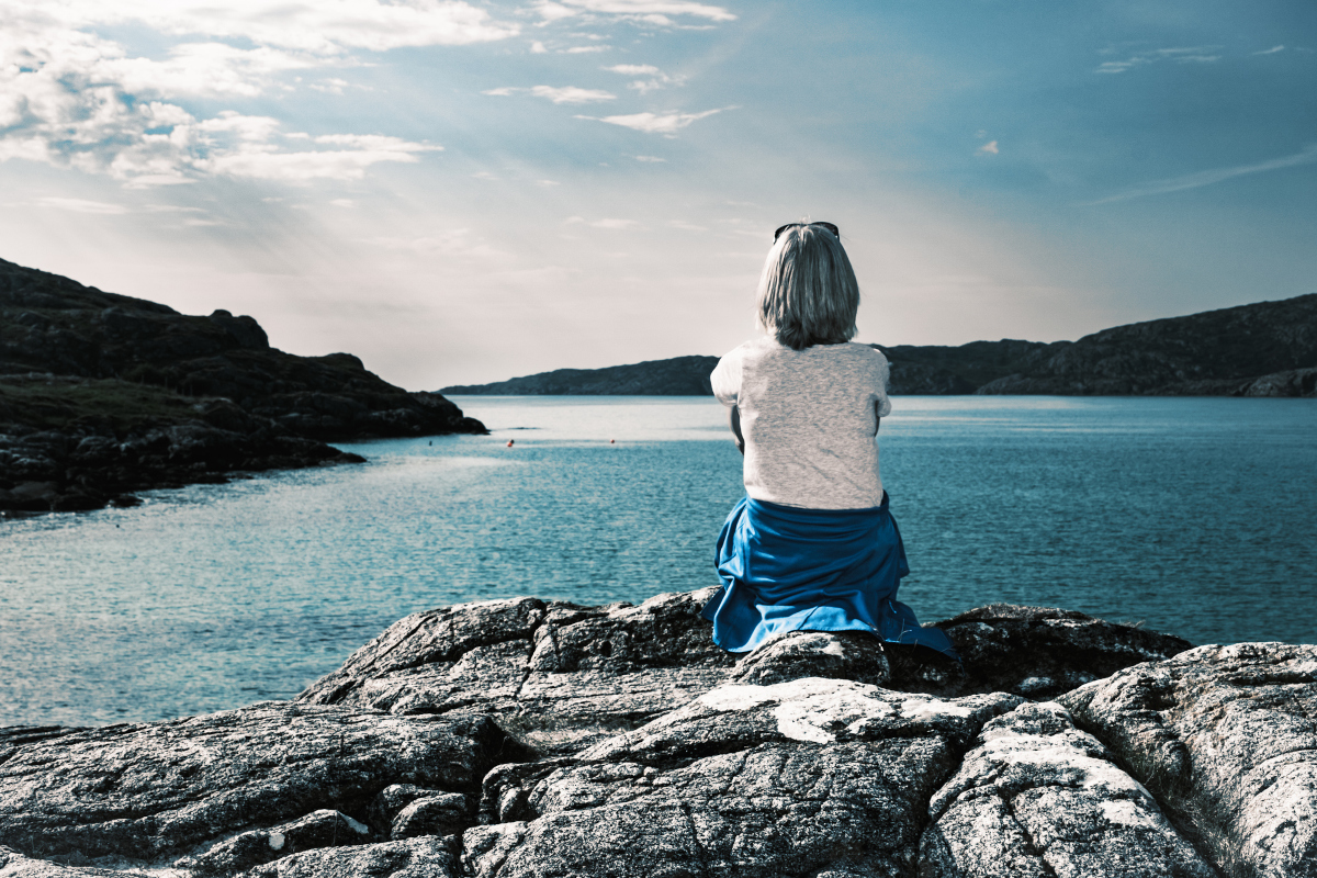 Woman sitting alone on rocky shoreline looking out over calm water, reflecting emotional overwhelm, stress, and nervous system exhaustion